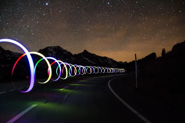 A long-exposure photo of a light trail formed as a horizontal spiral, above a road, in the middle of a starry night.